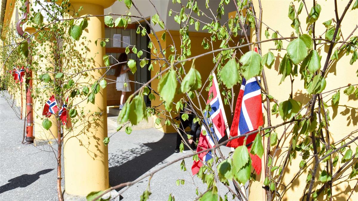 Foto av norskeflagg og bjørkepynt hengt opp på gule søyler - Klikk for stort bilete