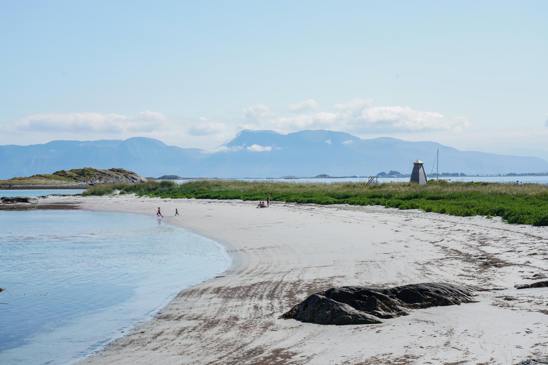 lang, brei, kvit sandstrand med roleg, blått hav og fjell i bakgrunnen