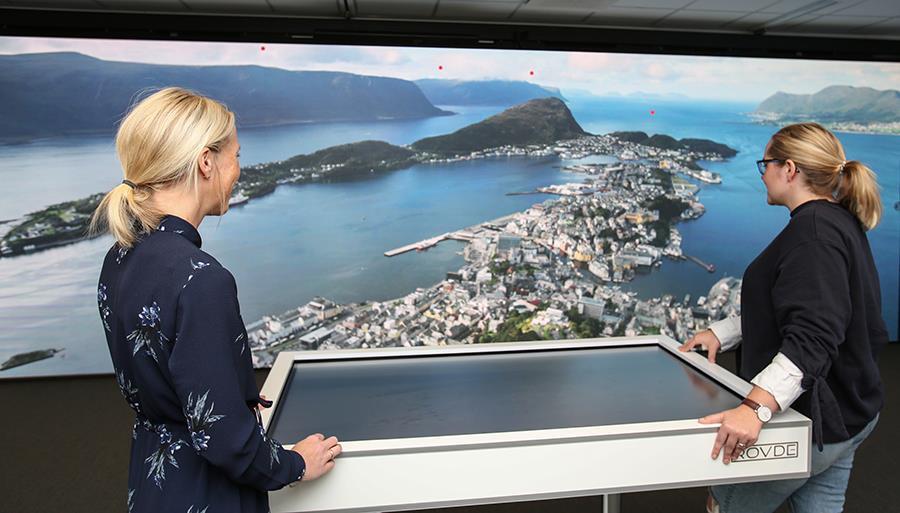 Ilustrasjon: To kvinner ser på et oversiktsbilde av Ålesund sentrum tatt fra Fjellstua, de står ved et bord der bordplata er en skjerm - Klikk for stort bilete