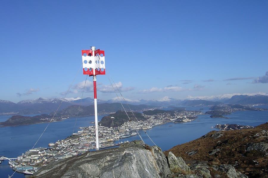 I forgrunnen et hvitt og rødt landemerke på en fjelltopp. Utsikt mot fjord og by i bakgrunnen. Fra Sukkertoppen mot Ålesund sentrum - Klikk for stort bilete